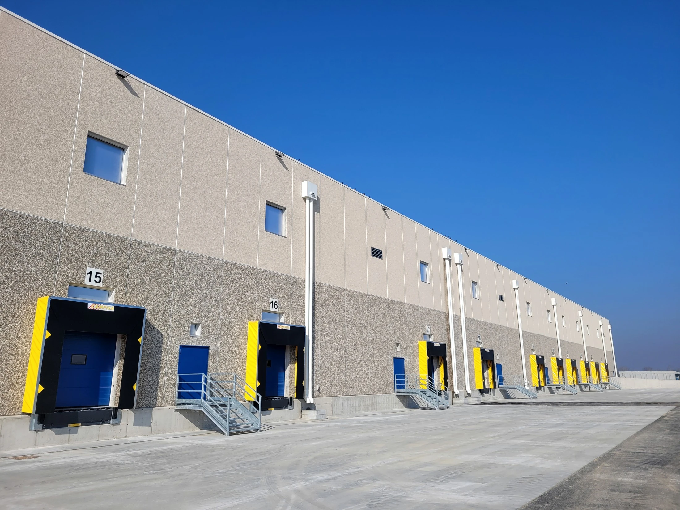 Facciata del magazzino con baie di carico numerate sotto un cielo azzurro terso, porte blu e gialle