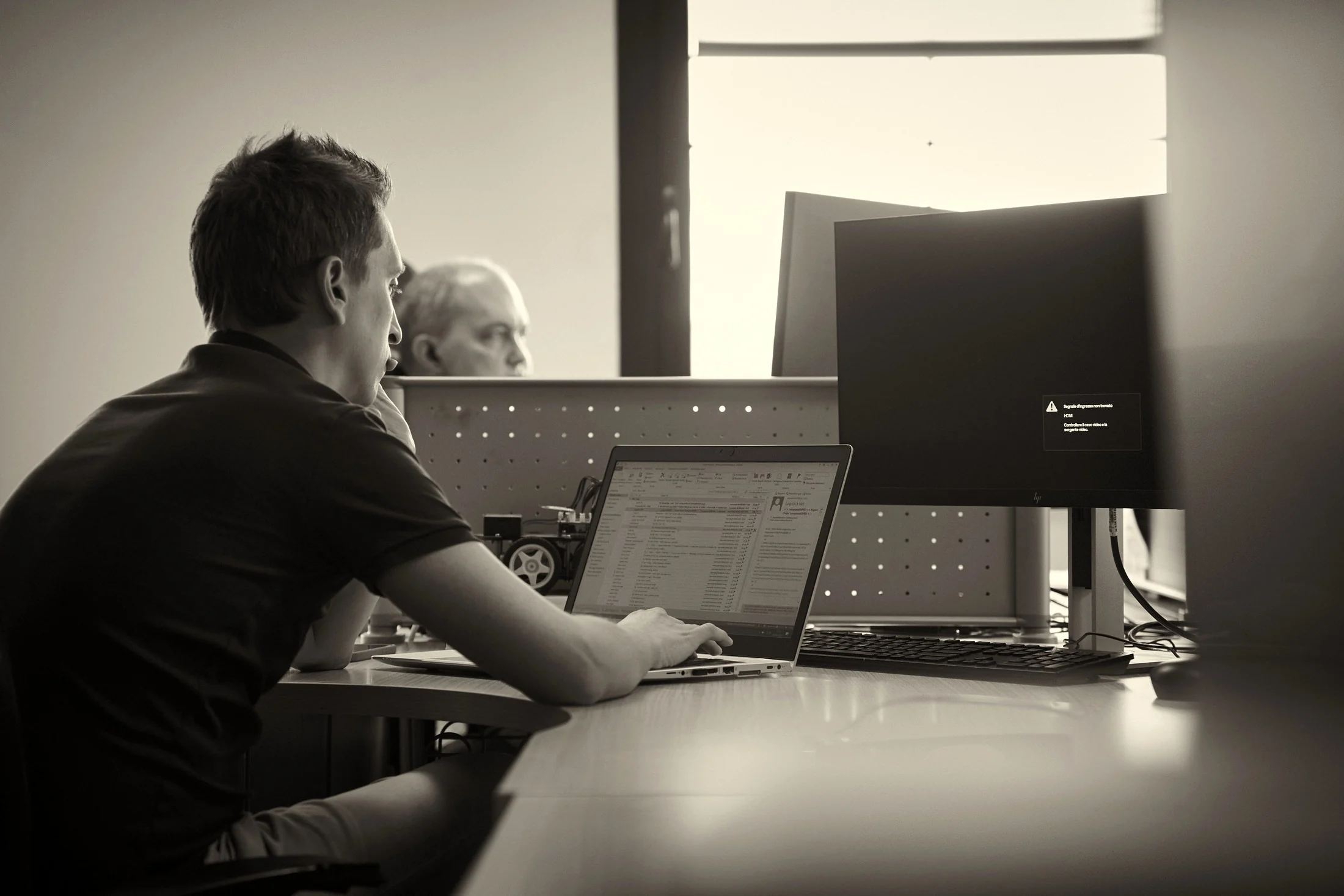 Persona al lavoro su un laptop con un collega a un secondo monitor sullo sfondo, bianco e nero